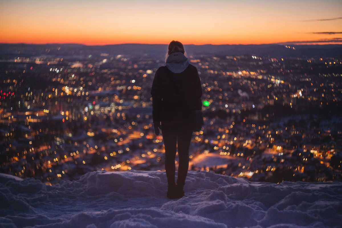 Eine Person steht im Schnee auf einem Hügel und blickt über die beleuchtete Stadt in der Dämmerung