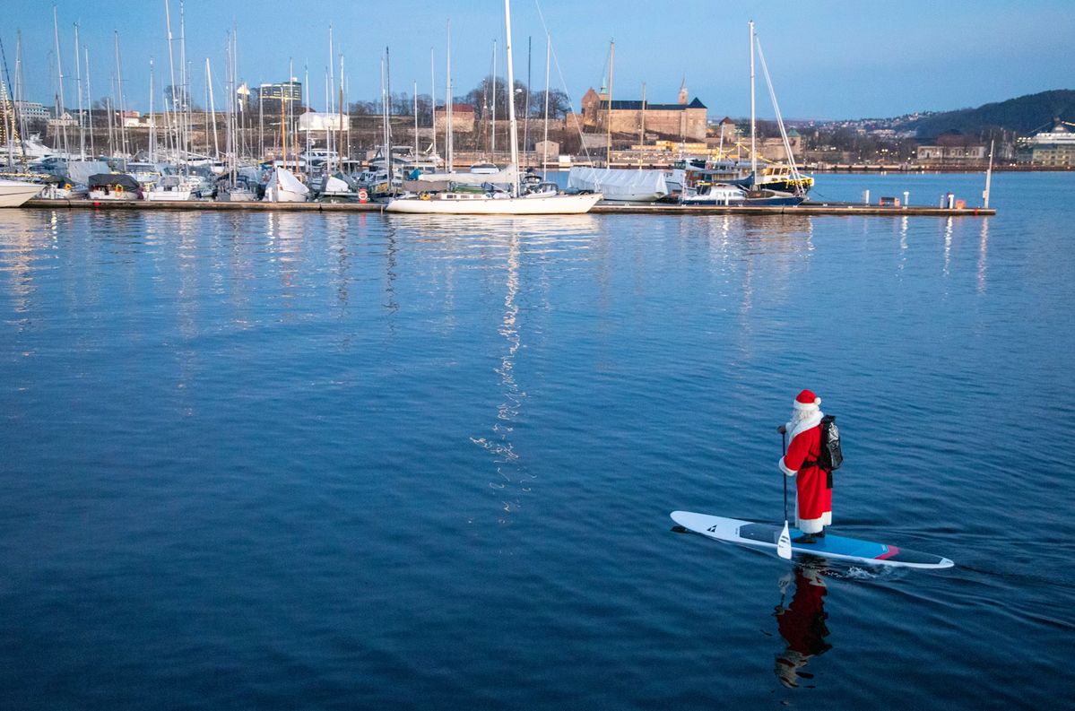 Ein Weihnachtsmann steht auf einem Stand-up-Paddle-Board im Hafen, dahinter Boote und die Festung Akershus